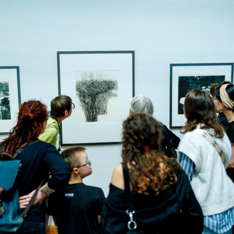 Muzeja apmeklētāju grupa stāv pie sienas un aplūko grafikas darbus rāmjos.