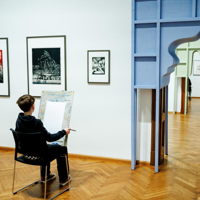 Zēns muzejā sēž pie molberta un zīmē, skatoties uz grafikas darbiem pie sienas blakus dekoratīvam durvju ailas atvērumam.