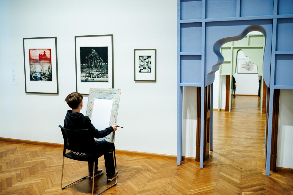 Zēns muzejā sēž uz krēsla un zīmē, skatoties uz pie sienas izstādītiem grafikas darbiem.