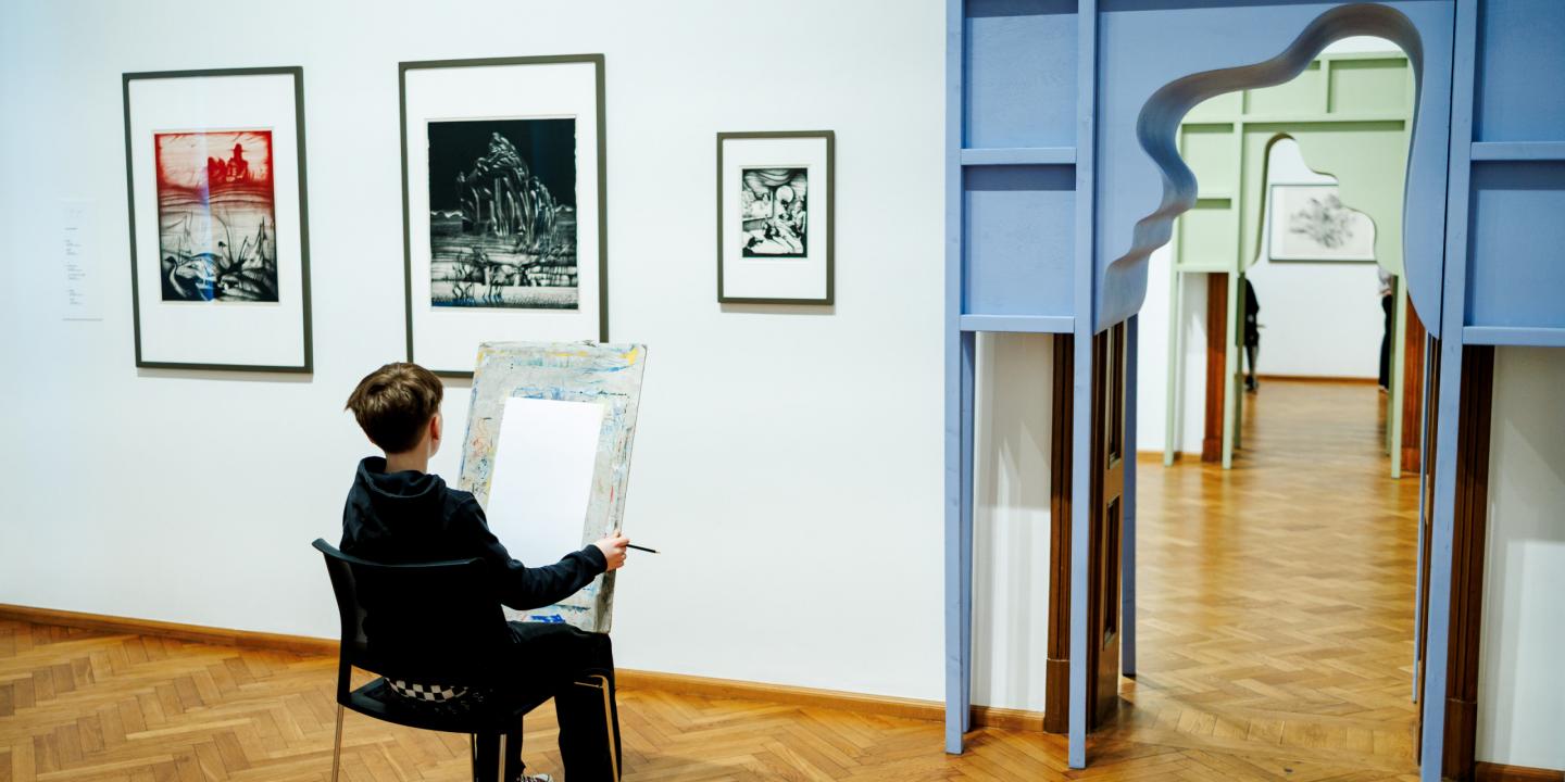Zēns muzejā sēž pie molberta un zīmē, skatoties uz grafikas darbiem pie sienas blakus dekoratīvam durvju ailas atvērumam.