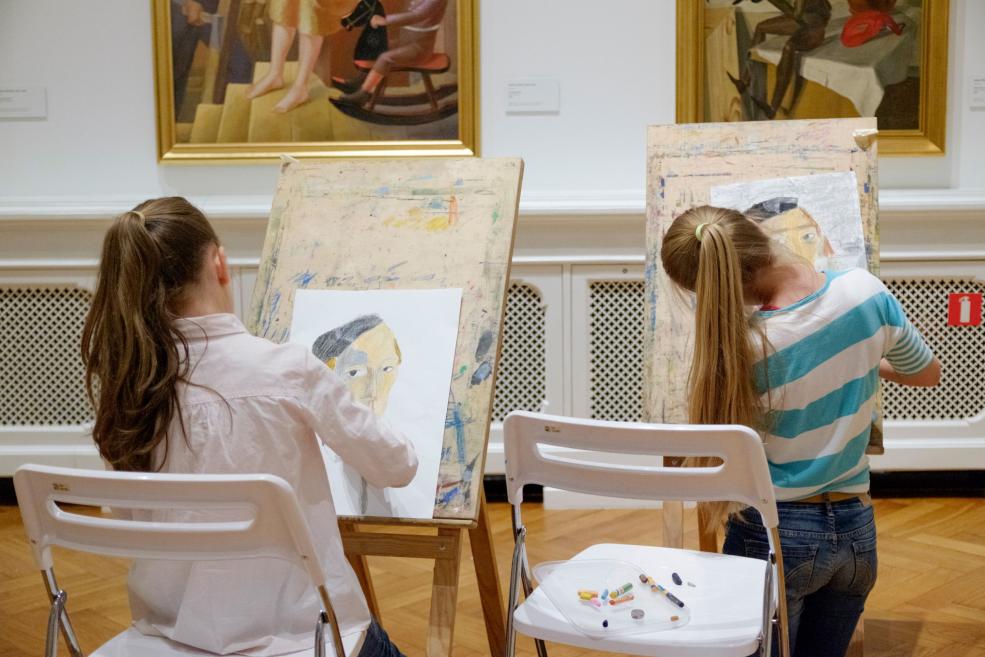 Divas meitenes muzejā pie molbertiem zīmē portretus, iedvesmojoties no gleznām pie sienas.