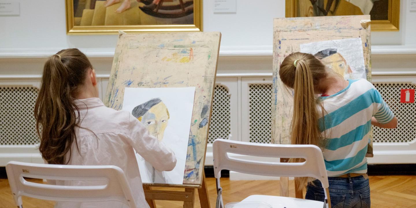 Divas meitenes muzejā sēž pie molbertiem un zīmē portretus, iedvesmojoties no gleznām pie sienas.