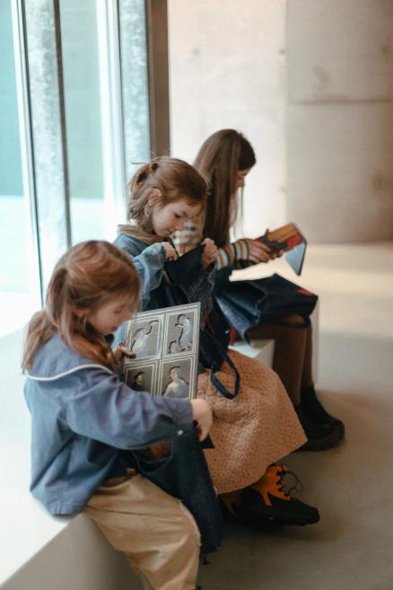 Trīs bērni muzejā sēž uz palodzes un aplūko katalogus, izņemot tos no somām.