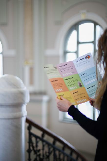 Apmeklētāja muzejā tur rokās krāsainu bukletu ar dažādām mākslas kategorijām.