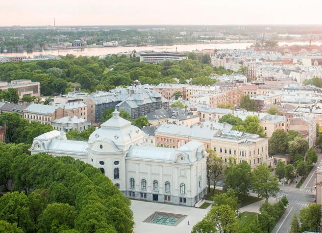 Skats no augšas uz pilsētas centru ar Latvijas Nacionālo mākslas muzeju, ko ieskauj koki, ielas un apbūve, fonā redzama upe.