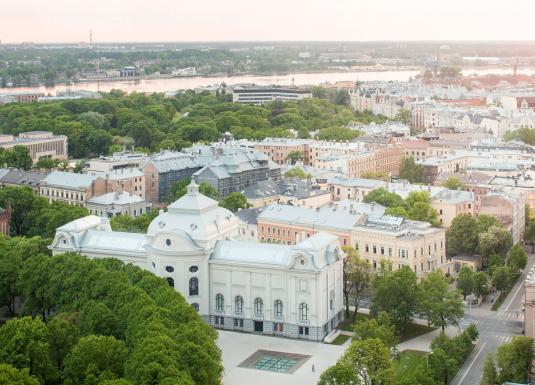 Skats no augšas uz pilsētas centru ar Latvijas Nacionālo mākslas muzeju, ko ieskauj koki, ielas un apbūve, fonā redzama upe.