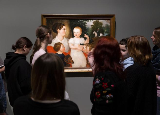 Apmeklētāju grupa muzejā stāv pie sienas un aplūko gleznu ar sievieti un bērniem.