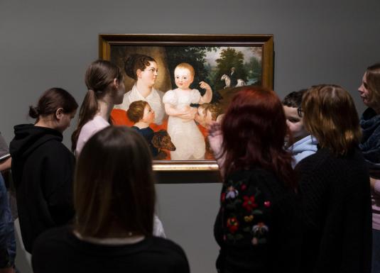 Apmeklētāju grupa muzejā stāv pie sienas un aplūko gleznu ar sievieti un bērniem.