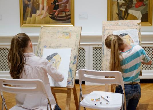 Divas meitenes muzejā sēž pie molbertiem un zīmē portretus, iedvesmojoties no gleznām pie sienas.