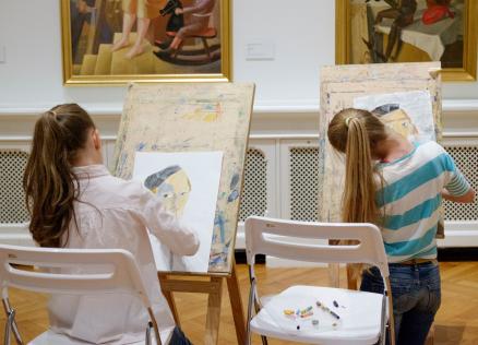 Divas meitenes muzejā sēž pie molbertiem un zīmē portretus, iedvesmojoties no gleznām pie sienas.