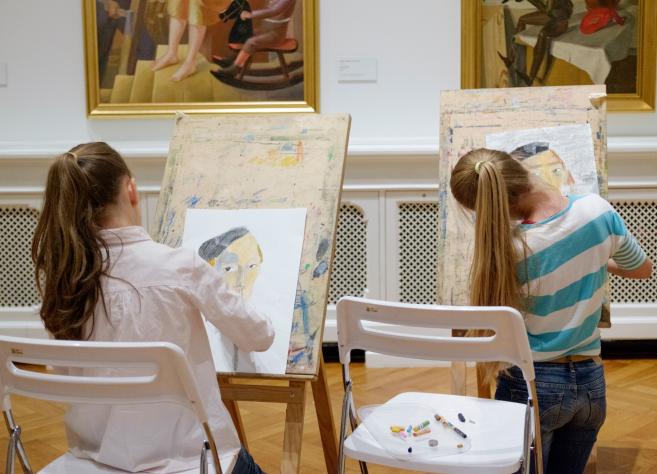 Divas meitenes muzejā sēž pie molbertiem un zīmē portretus, iedvesmojoties no gleznām pie sienas.