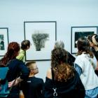 Muzeja apmeklētāju grupa stāv pie sienas un aplūko grafikas darbus rāmjos.