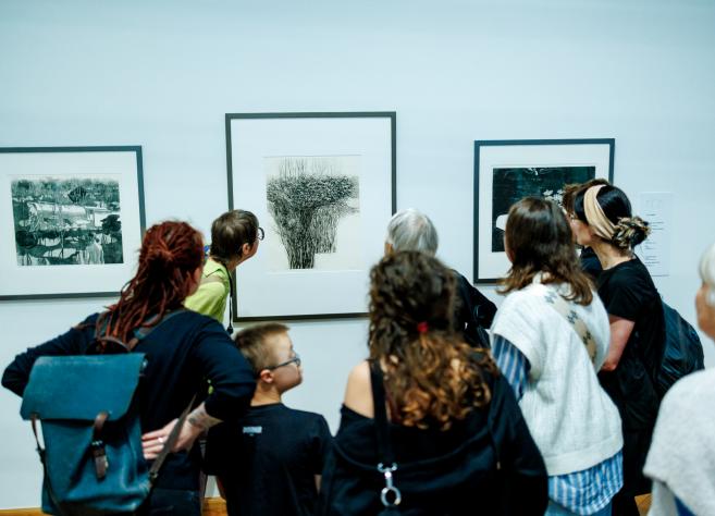 Muzeja apmeklētāju grupa stāv pie sienas un aplūko grafikas darbus rāmjos.