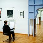 Zēns muzejā sēž pie molberta un zīmē, skatoties uz grafikas darbiem pie sienas blakus dekoratīvam durvju ailas atvērumam.