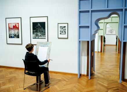 Zēns muzejā sēž pie molberta un zīmē, skatoties uz grafikas darbiem pie sienas blakus dekoratīvam durvju ailas atvērumam.