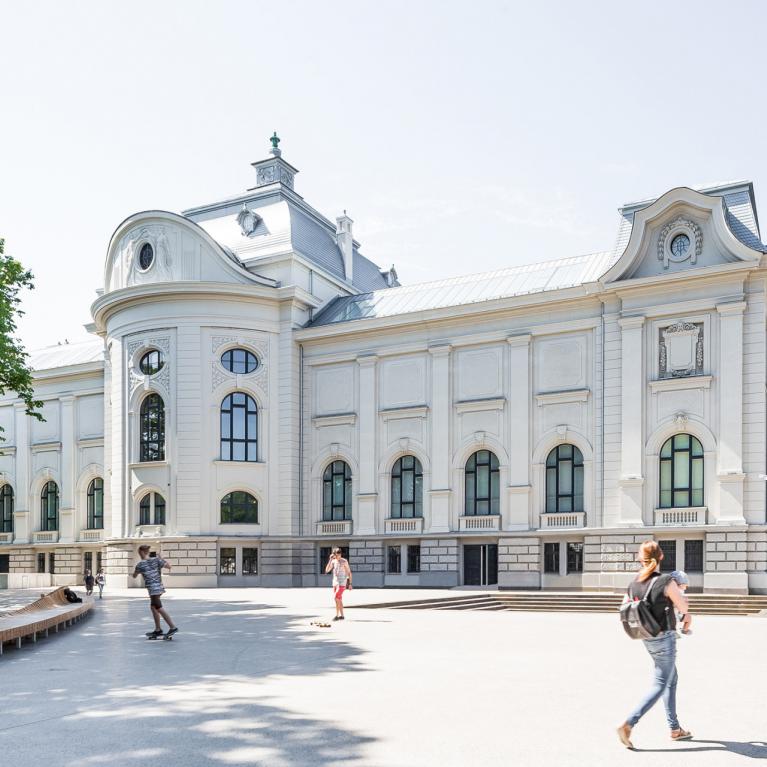 Gaiša, vēsturiska muzeja ēka parkā, priekšā redzami cilvēki, kas pastaigājas un atpūšas.