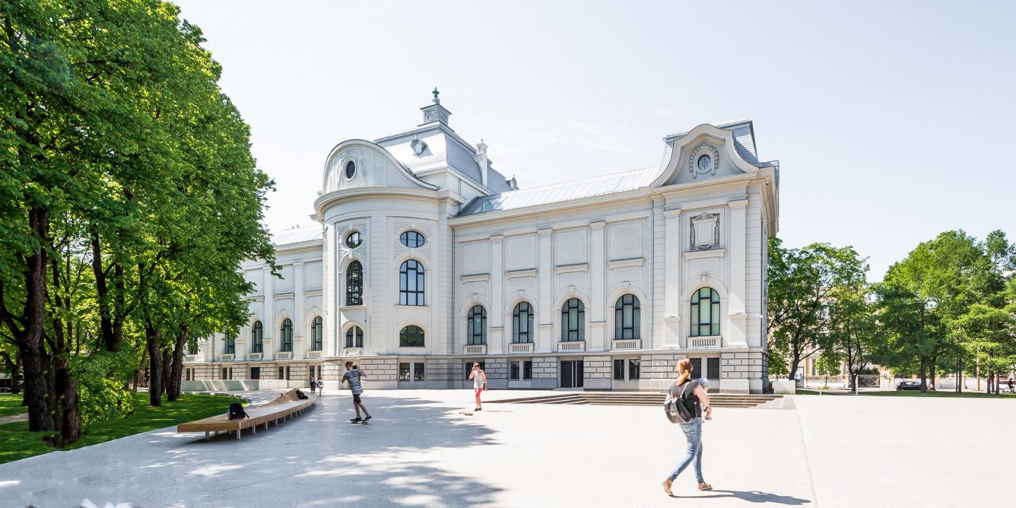 Gaiša, vēsturiska muzeja ēka parkā, priekšā redzami cilvēki, kas pastaigājas un atpūšas.