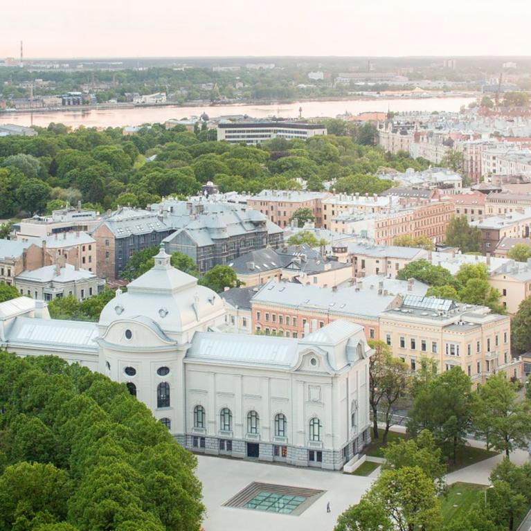 Skats no augšas uz pilsētas centru ar Latvijas Nacionālo mākslas muzeju, ko ieskauj koki, ielas un apbūve, fonā redzama upe.