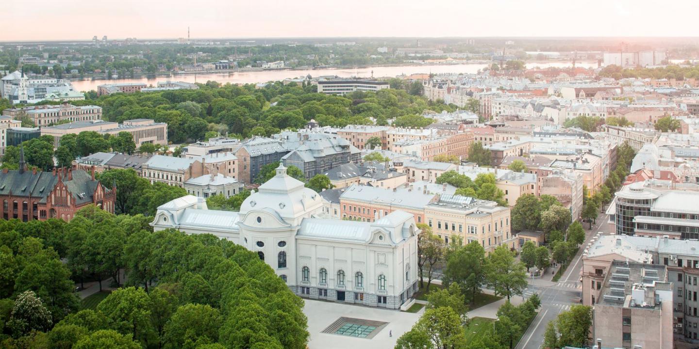 Skats no augšas uz pilsētas centru ar Latvijas Nacionālo mākslas muzeju, ko ieskauj koki, ielas un apbūve, fonā redzama upe.