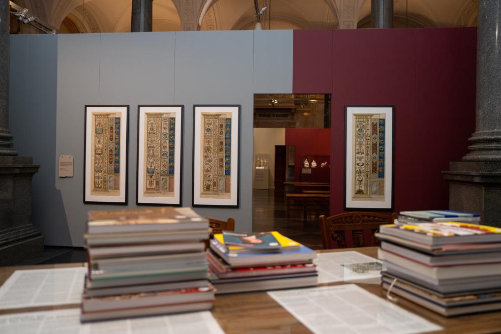 In a museum exhibition hall with gray and burgundy wall panels, four framed vertical decorative ornament drawings are displayed, with stacks of books on a table in the foreground.