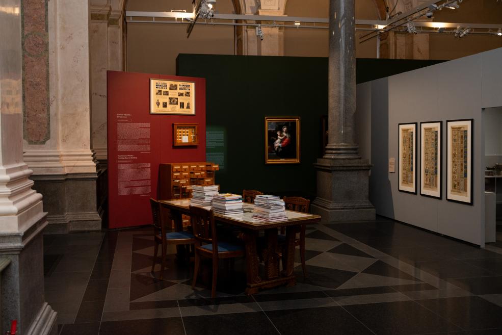 Museum exhibition hall with display panels, a framed painting, framed works on the wall, and a table covered with books.