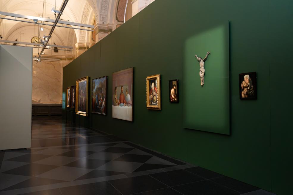 In a museum hall with a green partition wall, a row of paintings and a small crucifix sculpture are displayed on the wall.