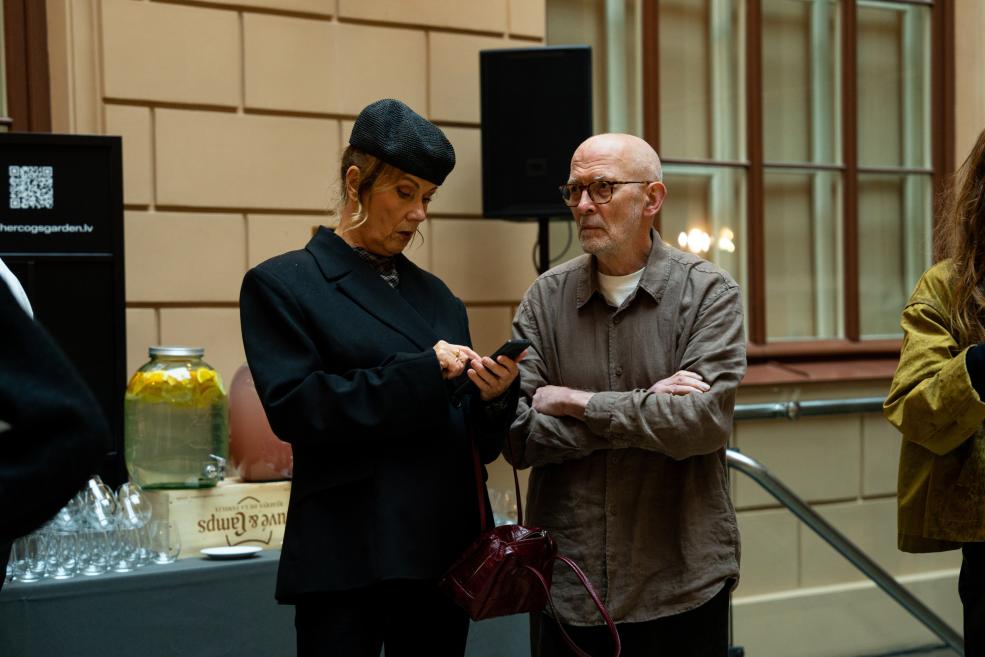 Two people stand indoors by a table with glasses and a jar, a woman in a black coat and hat looks at a smartphone while a man wearing glasses watches it.