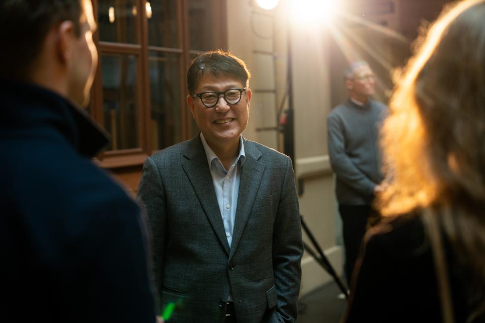 A smiling man wearing glasses and a gray suit stands indoors among other people, with bright light in the background.