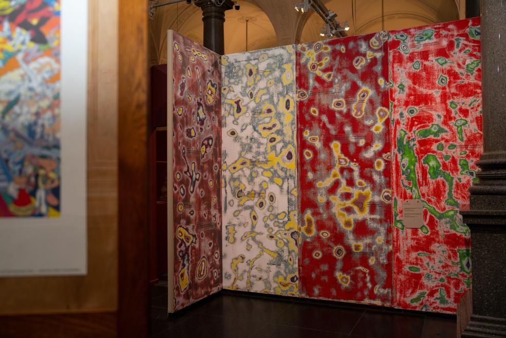 In a museum gallery, a large multi-panel abstract painting with red, white, gray, and green areas stands on folding screens.