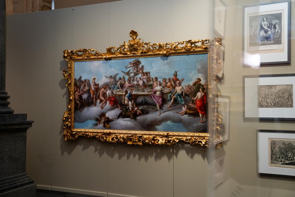 In a museum display, a large Baroque-style painting in an ornate gilded frame shows many figures among clouds, with several framed graphic works hanging on the wall to the right.
