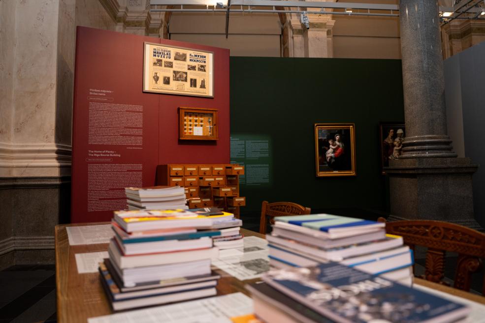 Museum interior display with a red and green wall, framed paintings, small display cases, and stacks of books on a table.