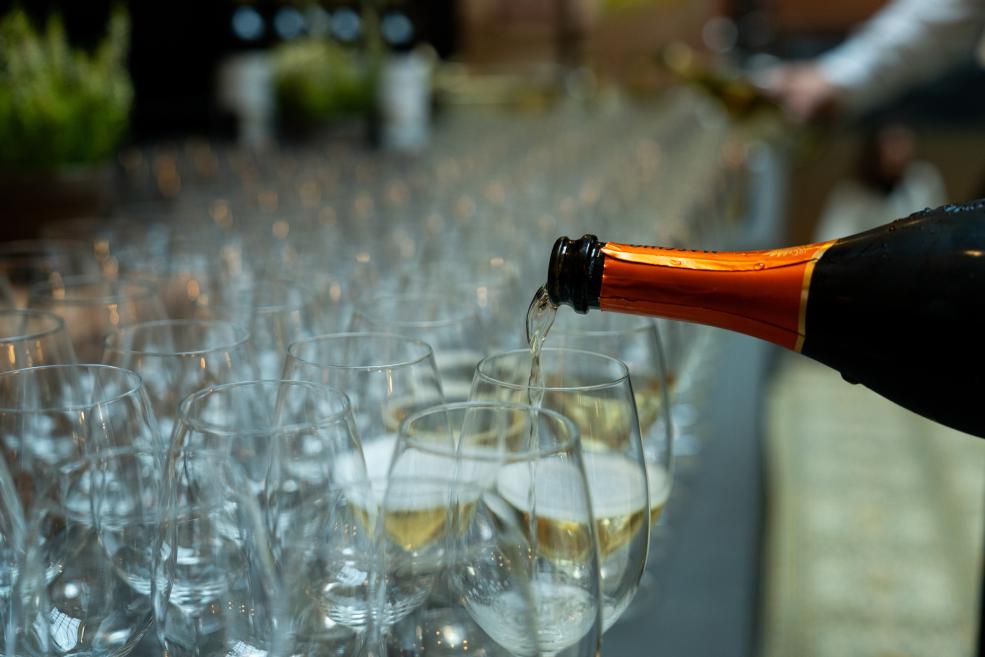 A sparkling wine bottle pours drink into rows of arranged stemmed glasses.