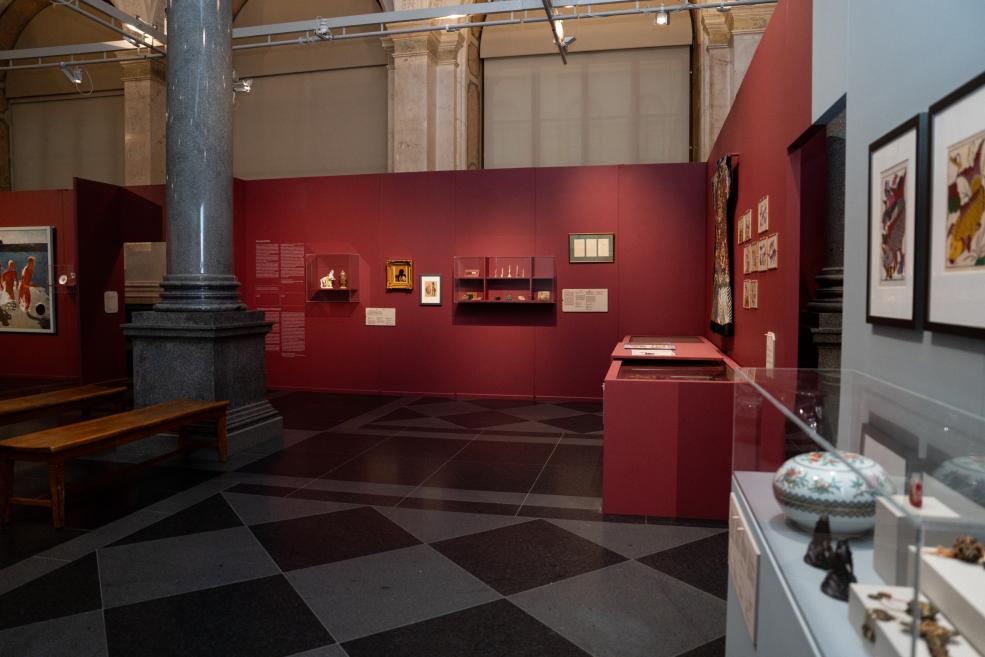 Museum exhibition hall with burgundy walls, a black-and-gray floor, columns, and artworks and objects displayed in cases and on the walls.