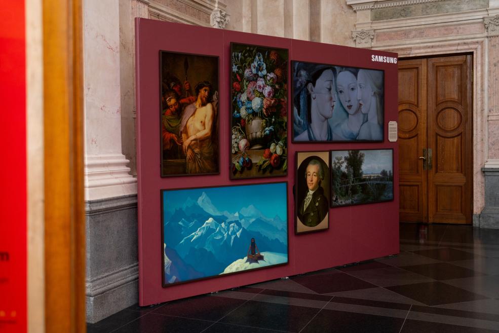 Several reproduction paintings, including portraits, a flower still life and a landscape, are displayed on a burgundy exhibition panel in a museum hall, with a Samsung logo on the panel.