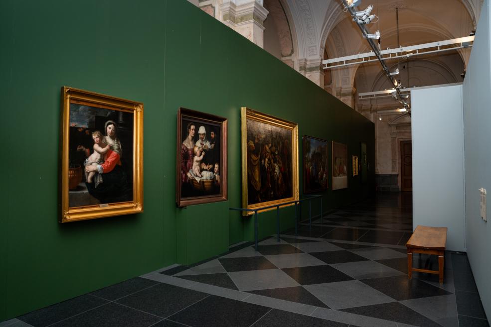 Several framed paintings are displayed on a green wall in a museum gallery, with high vaulted ceilings and a black-and-gray patterned floor in the background.