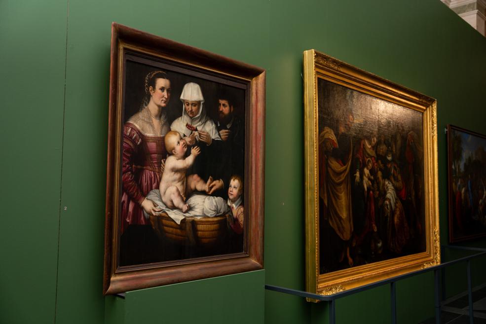 In a gallery hall, two large framed paintings hang on a green wall, with the left showing a woman, a nun and a child, and the right a dark religious scene.