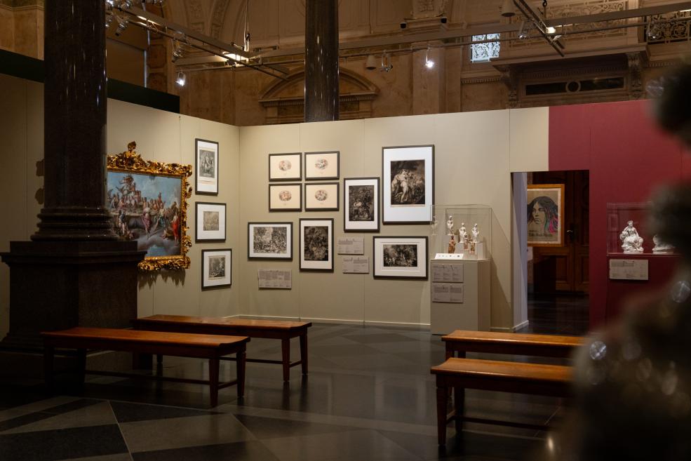 An art exhibition in a museum gallery shows framed prints, paintings, sculptures in display cases, and wooden benches in the foreground.