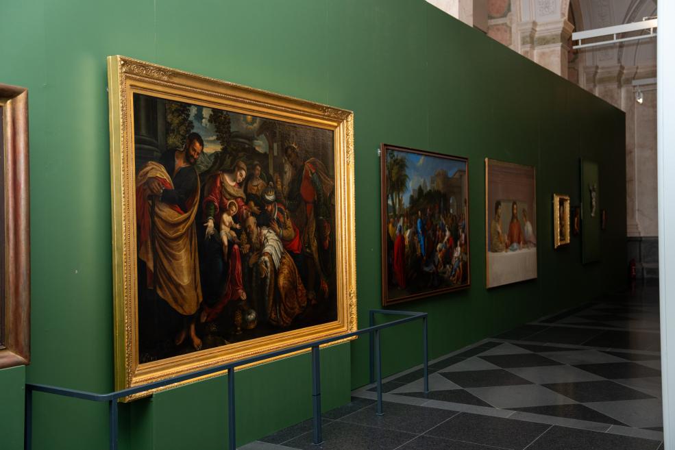 Several paintings are displayed in a row on a green wall in a museum gallery, including a large work in a gilded frame with a religious scene.