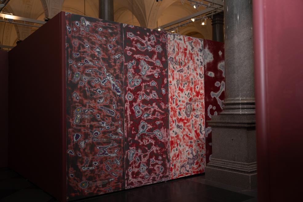 A large spatial installation with red-and-white abstract patterns on panels stands in a museum hall beside a stone column.