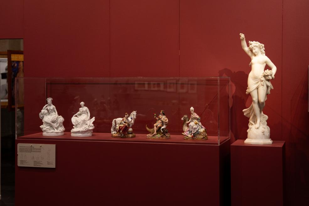 Several porcelain figurines, including a large female statue, displayed in a glass case and on a pedestal against a burgundy museum wall.