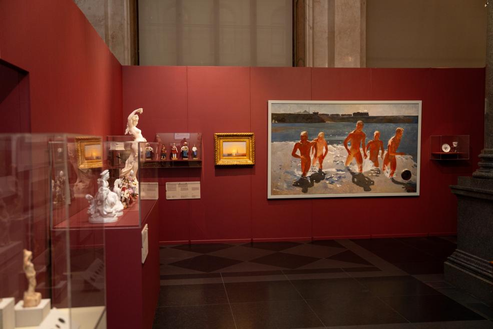 Museum exhibition hall with red walls, displaying sculptures, paintings and decorative objects in cases and on the wall, including a large painting of nude bathers by the sea.