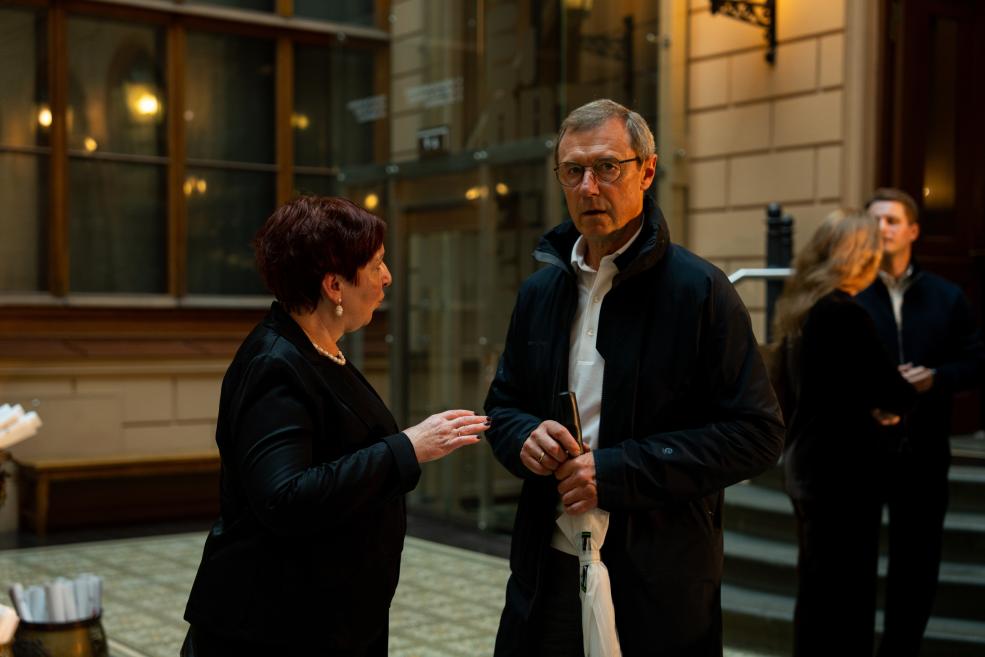 Two people in dark clothing converse in a bright museum foyer with a glass backdrop.
