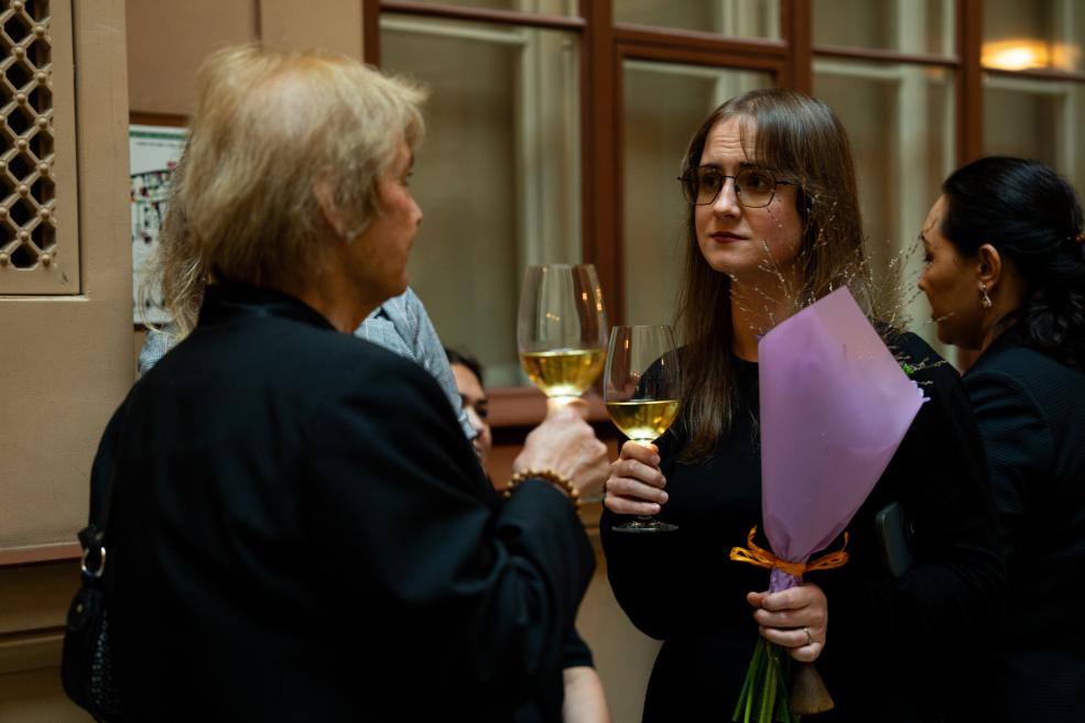 Three women in a room with large windows hold glasses of white wine, and the young woman with glasses holds a purple-wrapped bouquet of flowers.