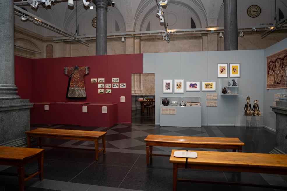 In a museum gallery with high arched ceilings and columns, an exhibition displays a kimono, small framed works, ceramics and sculptures, with wooden benches in the foreground.