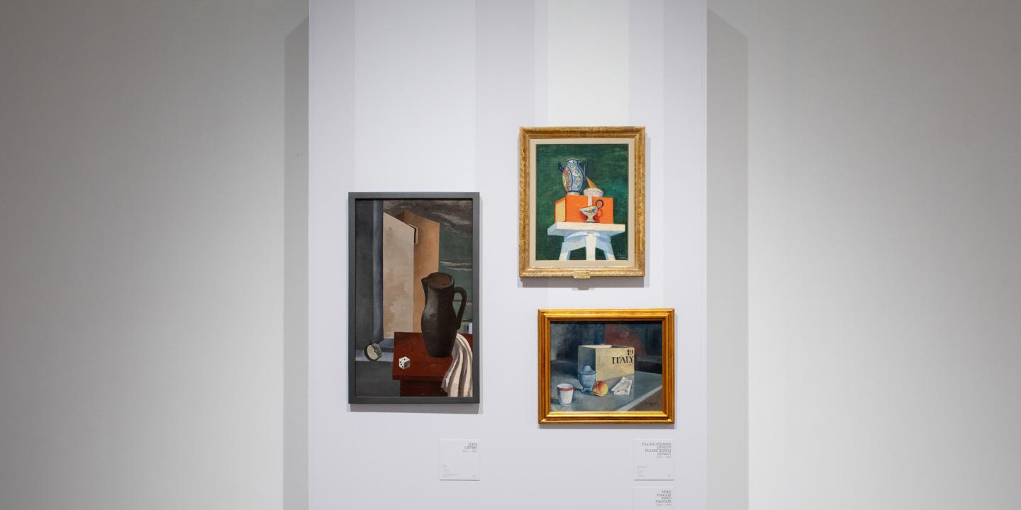 Trīs klusās dabas gleznas izstādē uz baltas sienas – ar krūzi, traukiem un grāmatu kompozīcijām.