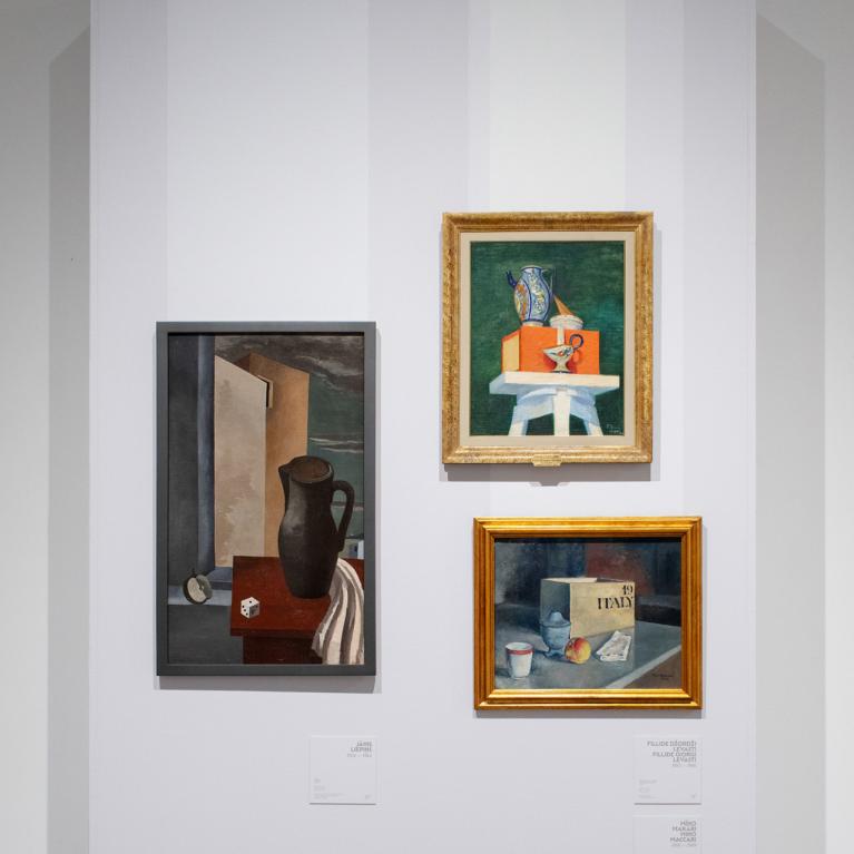 Trīs klusās dabas gleznas izstādē uz baltas sienas – ar krūzi, traukiem un grāmatu kompozīcijām.