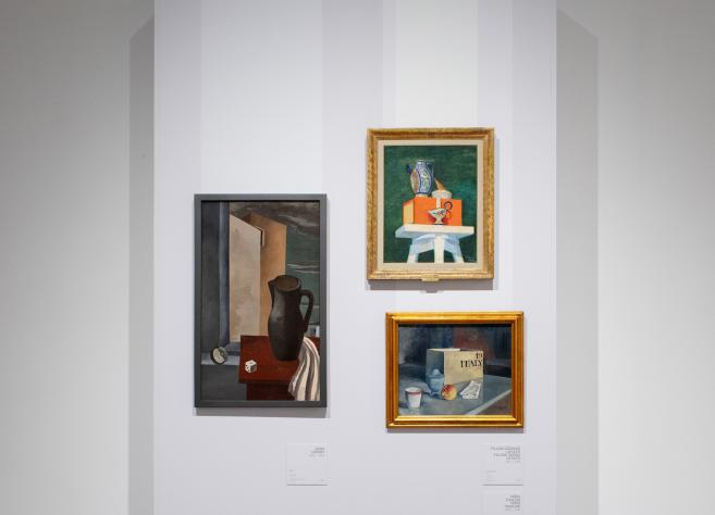 Trīs klusās dabas gleznas izstādē uz baltas sienas – ar krūzi, traukiem un grāmatu kompozīcijām.