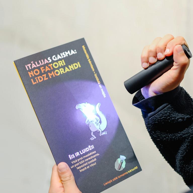 Apmeklētājs ar lukturīti izgaismo bukletu “Itālijas gaisma: no Fatori līdz Morandi”, kur redzams paslēpts ilustrēts tēls.
