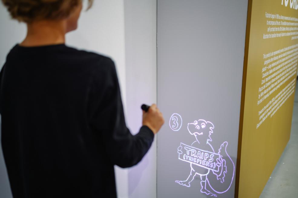 Bērns ar lukturīti izgaismo ilustrāciju ar tēlu Luidži pie teksta sienas izstādē.