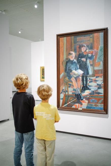 Divi bērni stāv LNMM izstāžu zālē un skatās uz lielu gleznu, kur attēloti muzicējoši bērni.